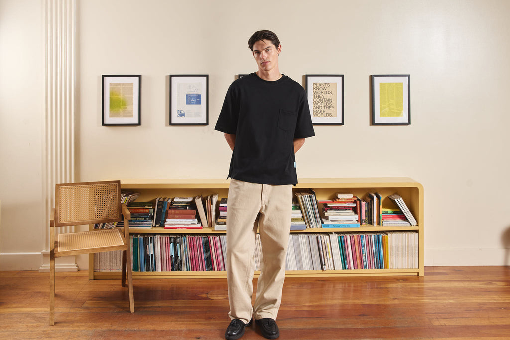 Man standing in a room with a bookshelf filled with books and framed pictures on the wall.